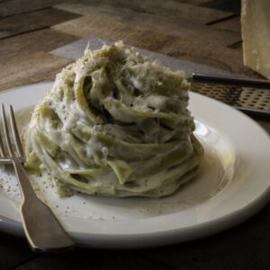 Swirl of spinach fettuccine on a plate with fork and pecorino cheese in the background.