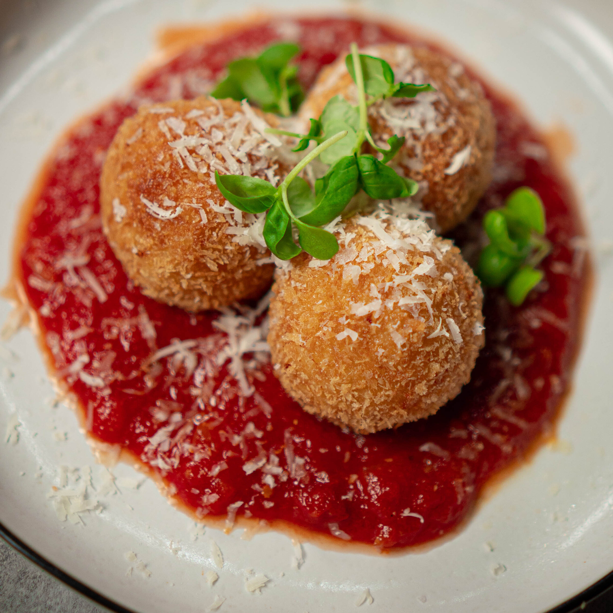 3 fried arancini on a puddle of marinara sauce on a white plate garnished with grated cheese and micro basil.