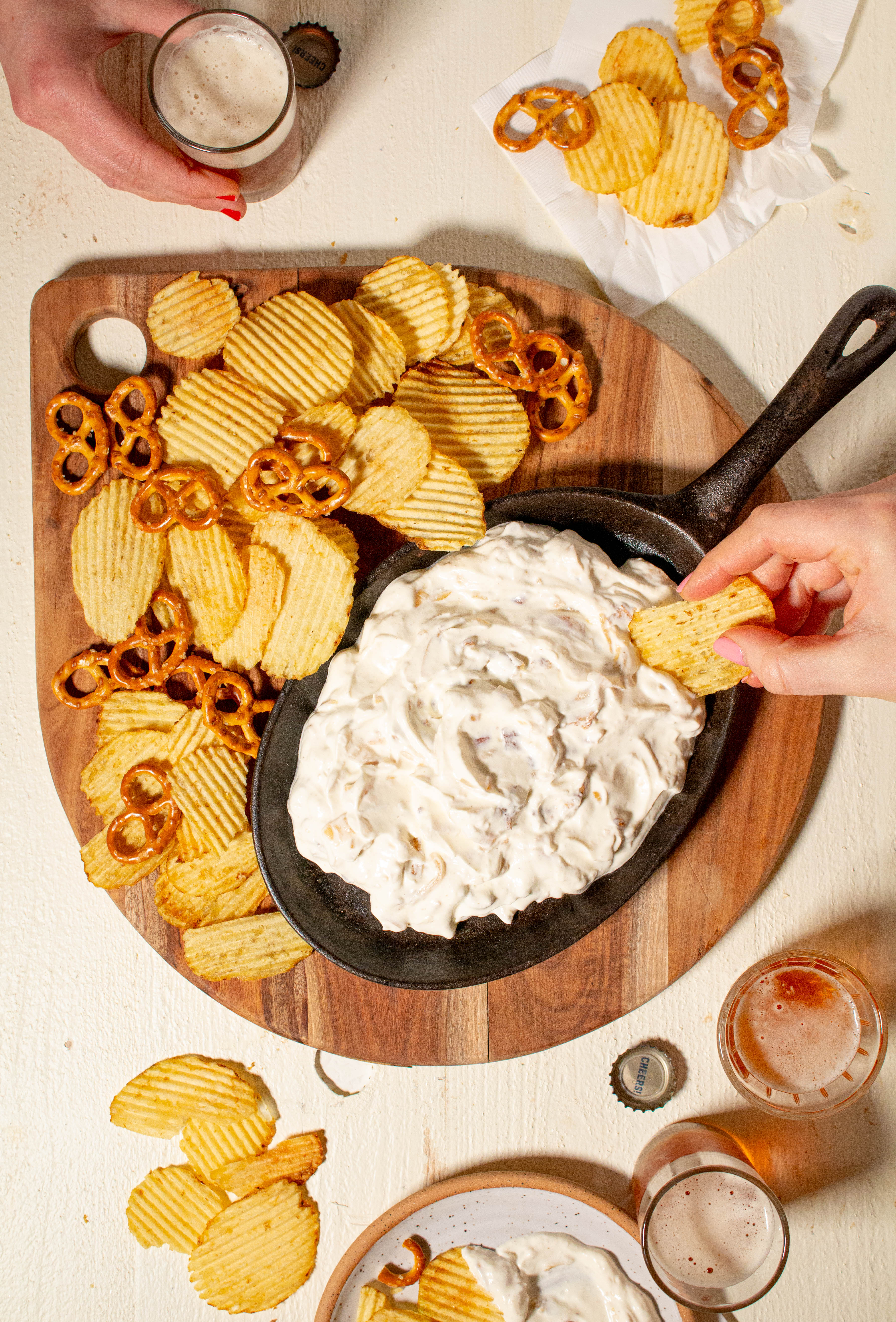 Cutting board with oval cast-iron pan with swirls of dip surrounded by chips, mini pretzels and beer.
