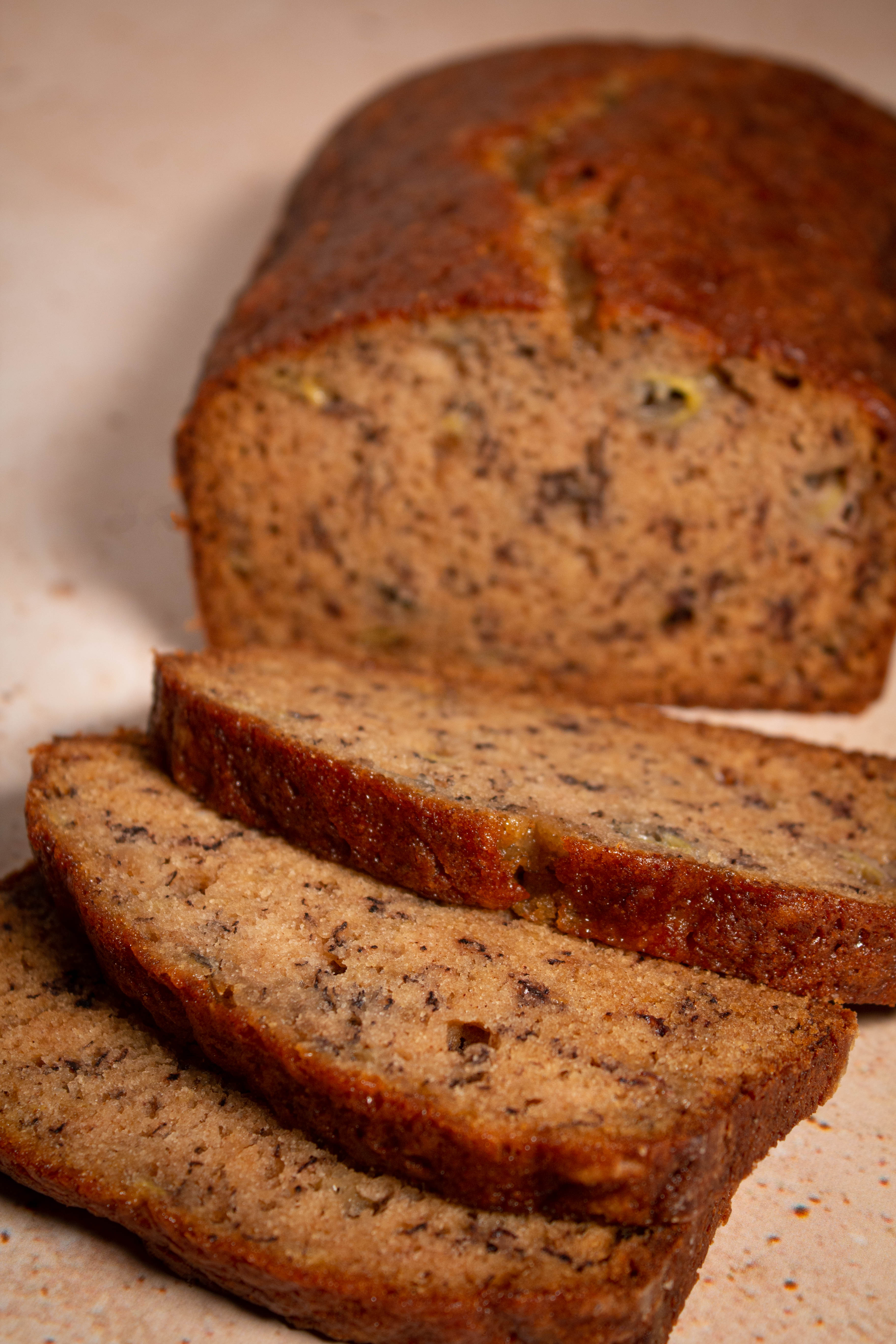 Three slices of shingled banana bread with remaining loaf in the background on a speckled, sand-colored surface