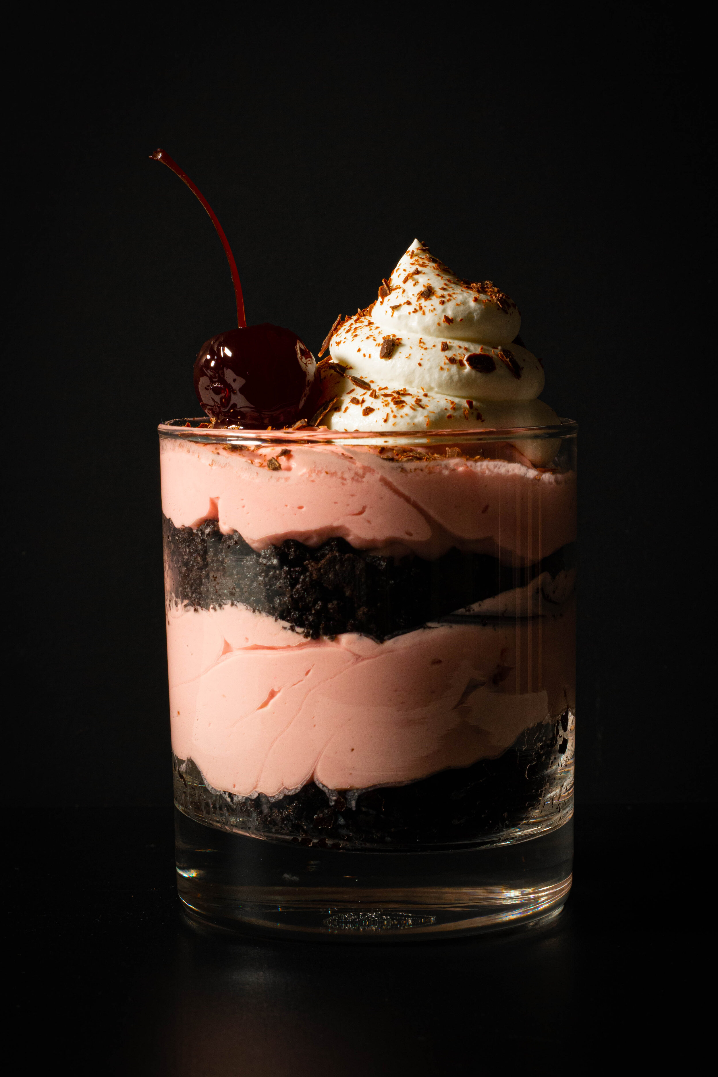 Parfait with layers of pink cream cheese filling and chocolate cookie crumbles in a rocks glass with whipped cream garnish and a cherry.