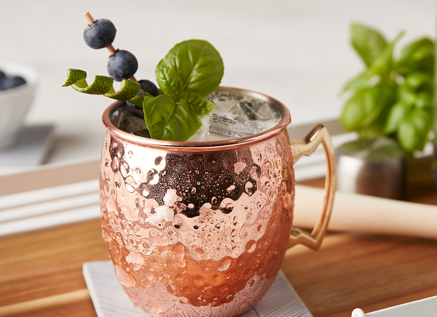 Copper mule glass with condensation, filled with a clear effervescent cocktail with mint, blueberry and lime peel garnish.