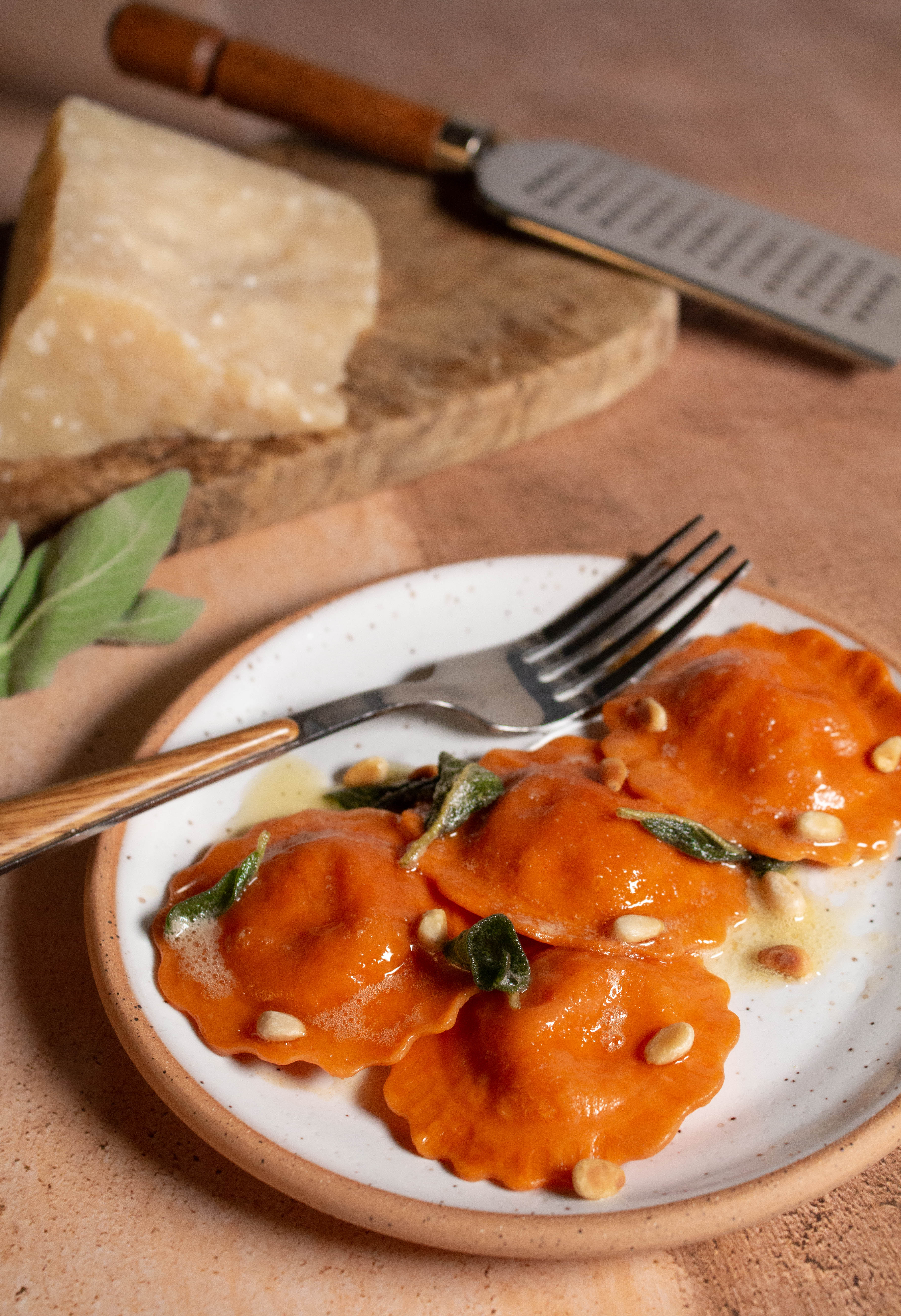 Butternut squash ravioli with brown butter sage sauce on a white plate with block of cheese and grater in the background.