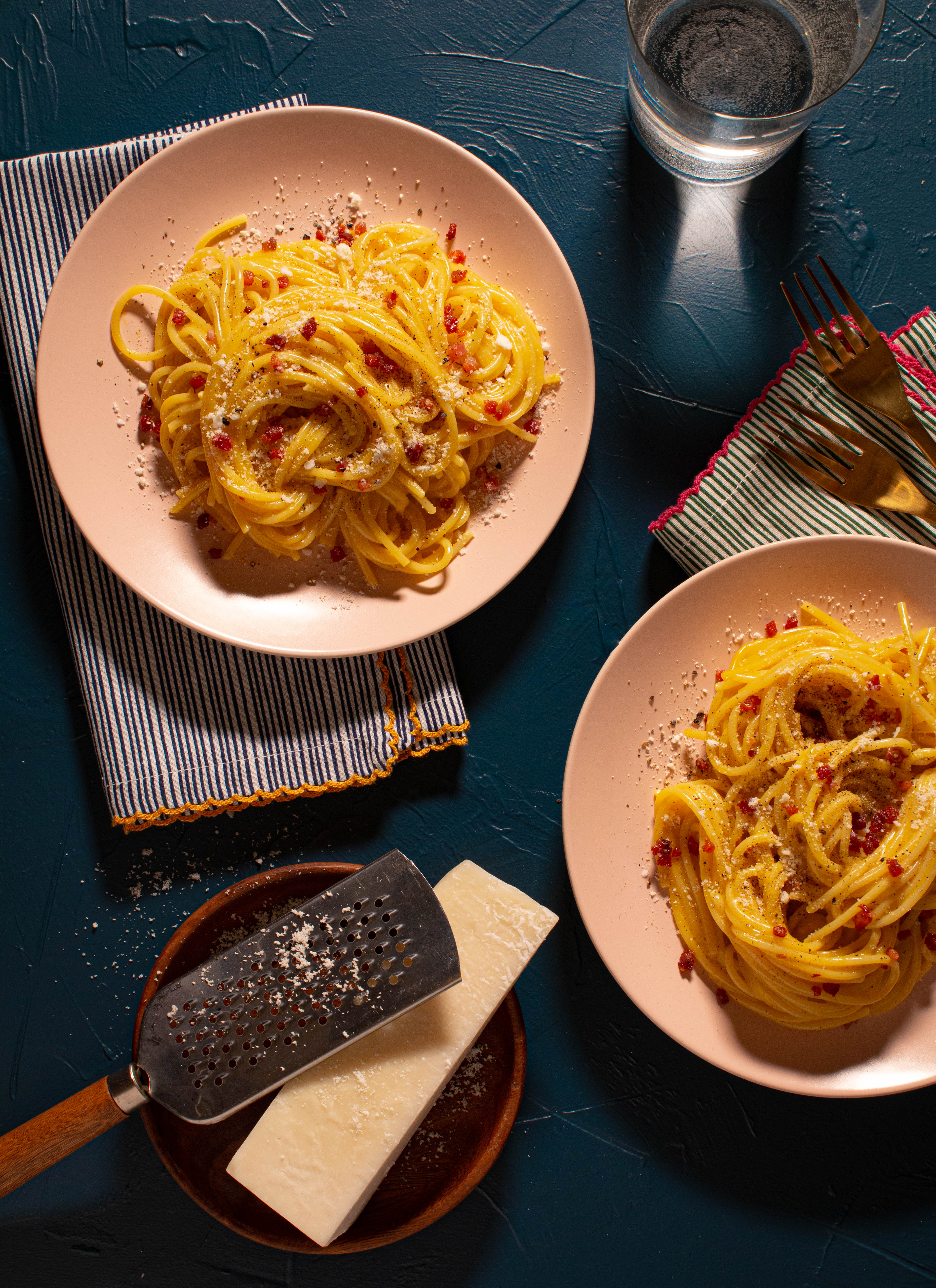 Swirls of spaghetti carbonara on blush pink plates atop a navy surface with a wedge of pecorino and grater.