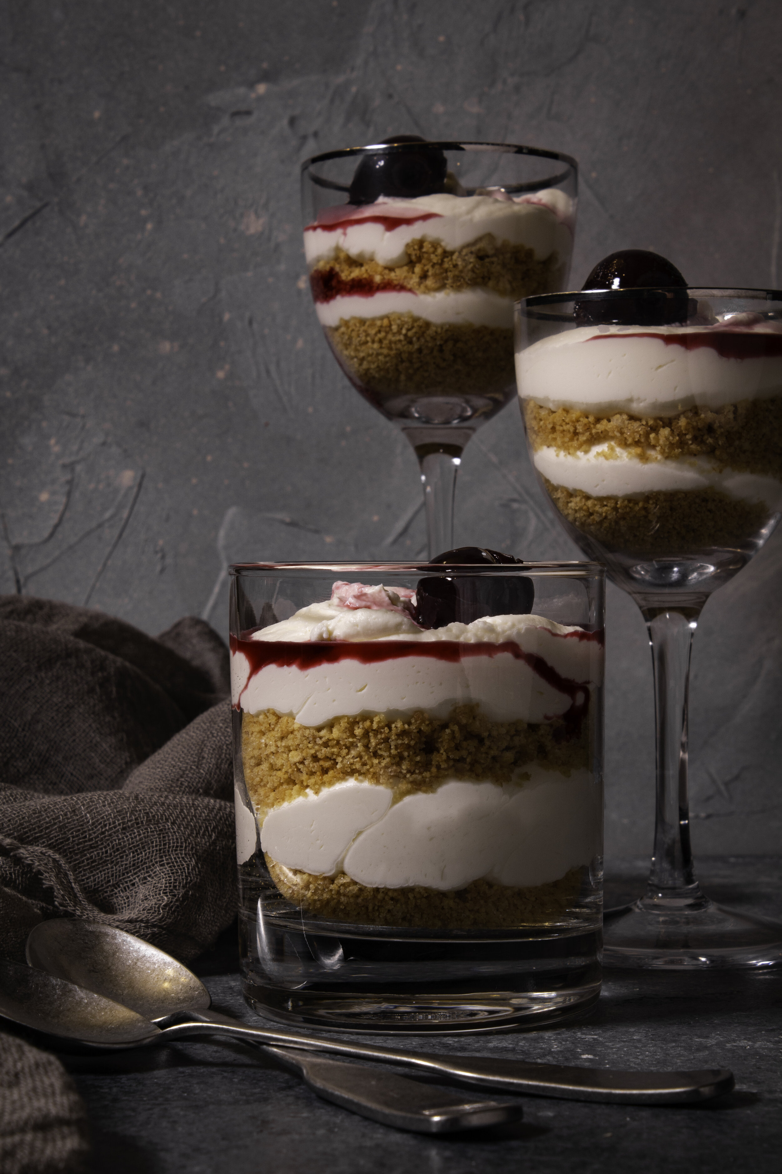 Various sized cups filled with layers of graham crumble, cheesecake mouse and Amarena cherries with a speckled gray background.