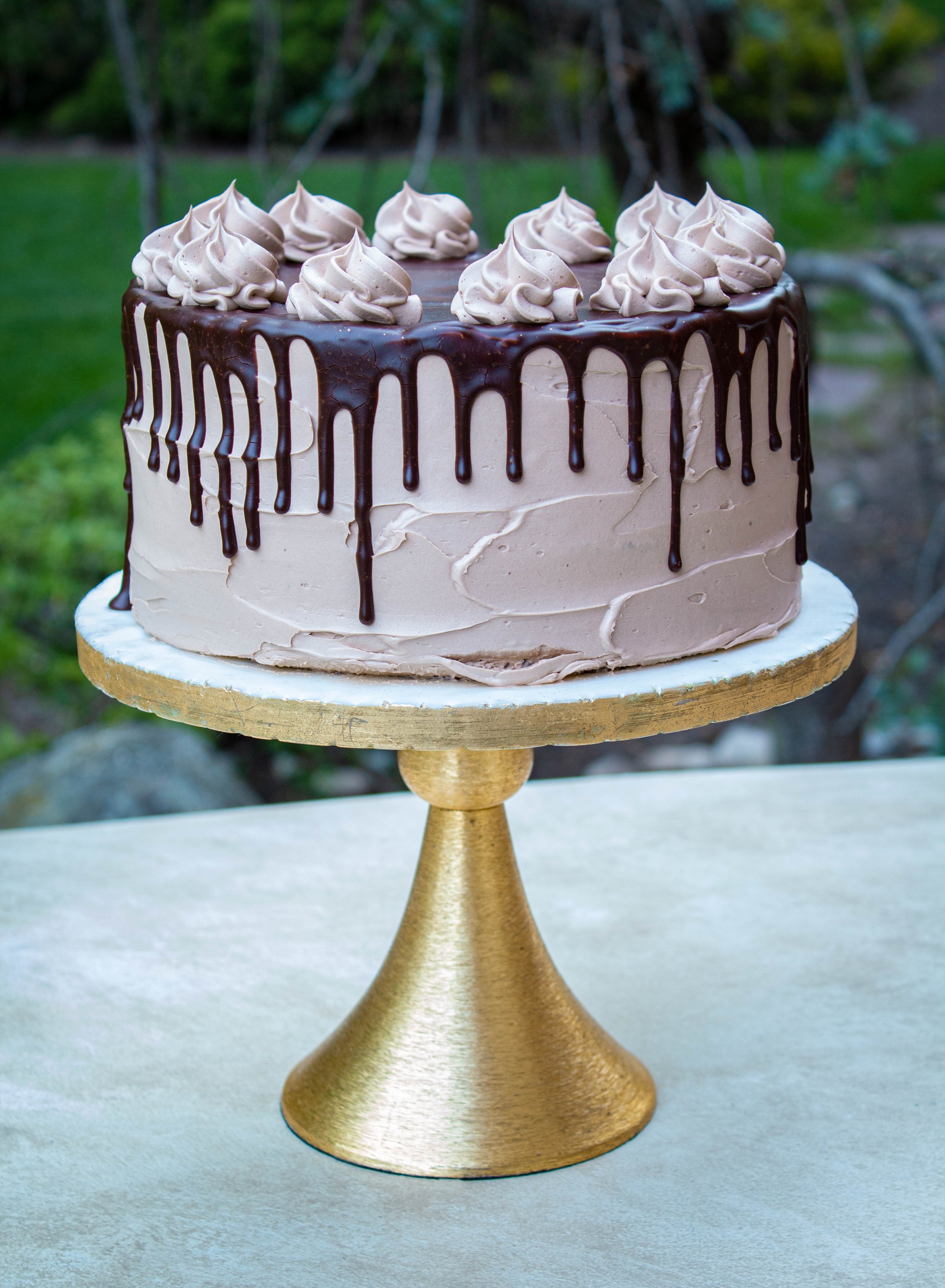 Chocolate Cake with Chocolate Glaze Drizzle on a gold and white marble cake stand in an outdoor setting.