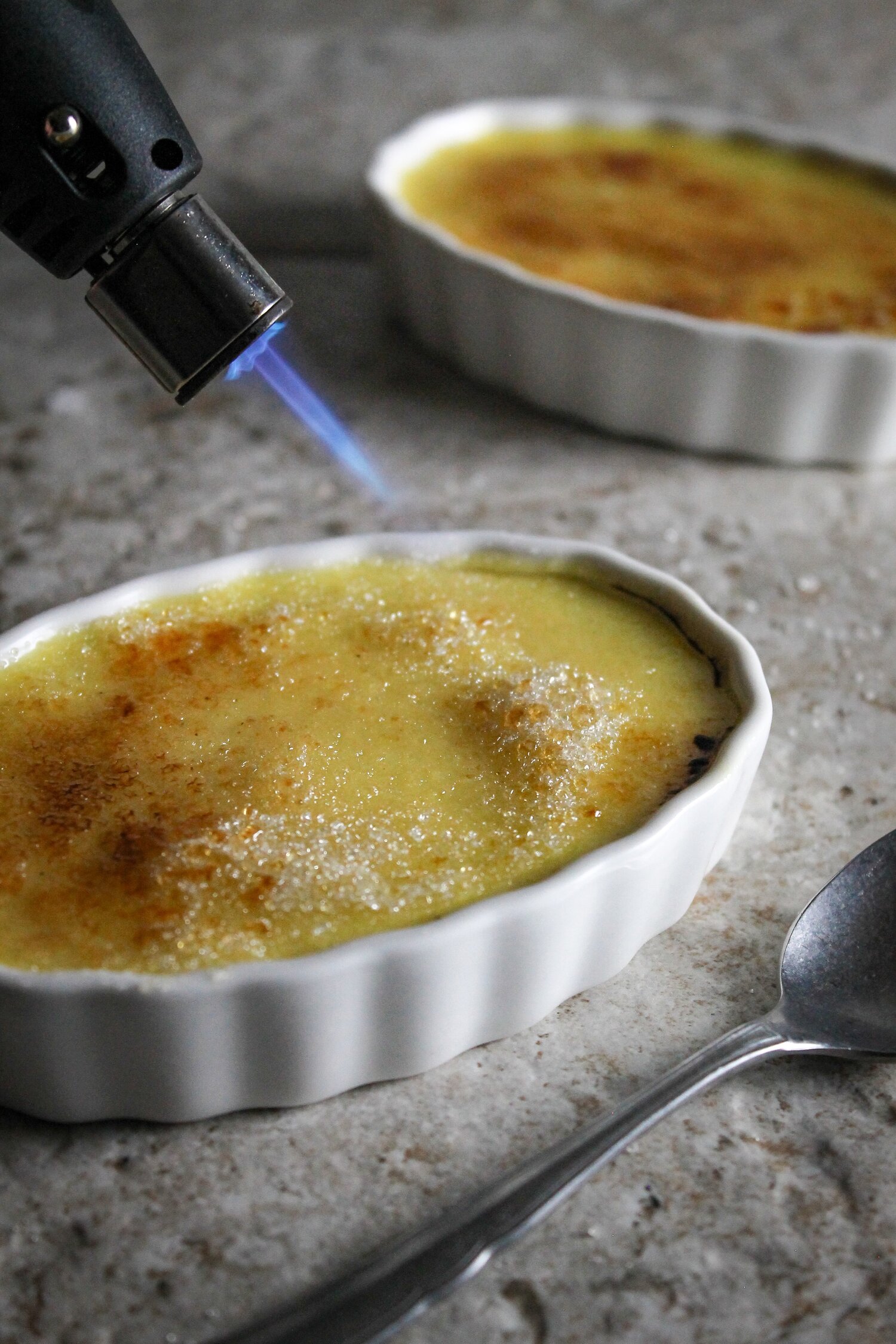 Torch caramelizing sugar on a ramekin with set creme brûlée with a matte silver spoon in the foreground. 