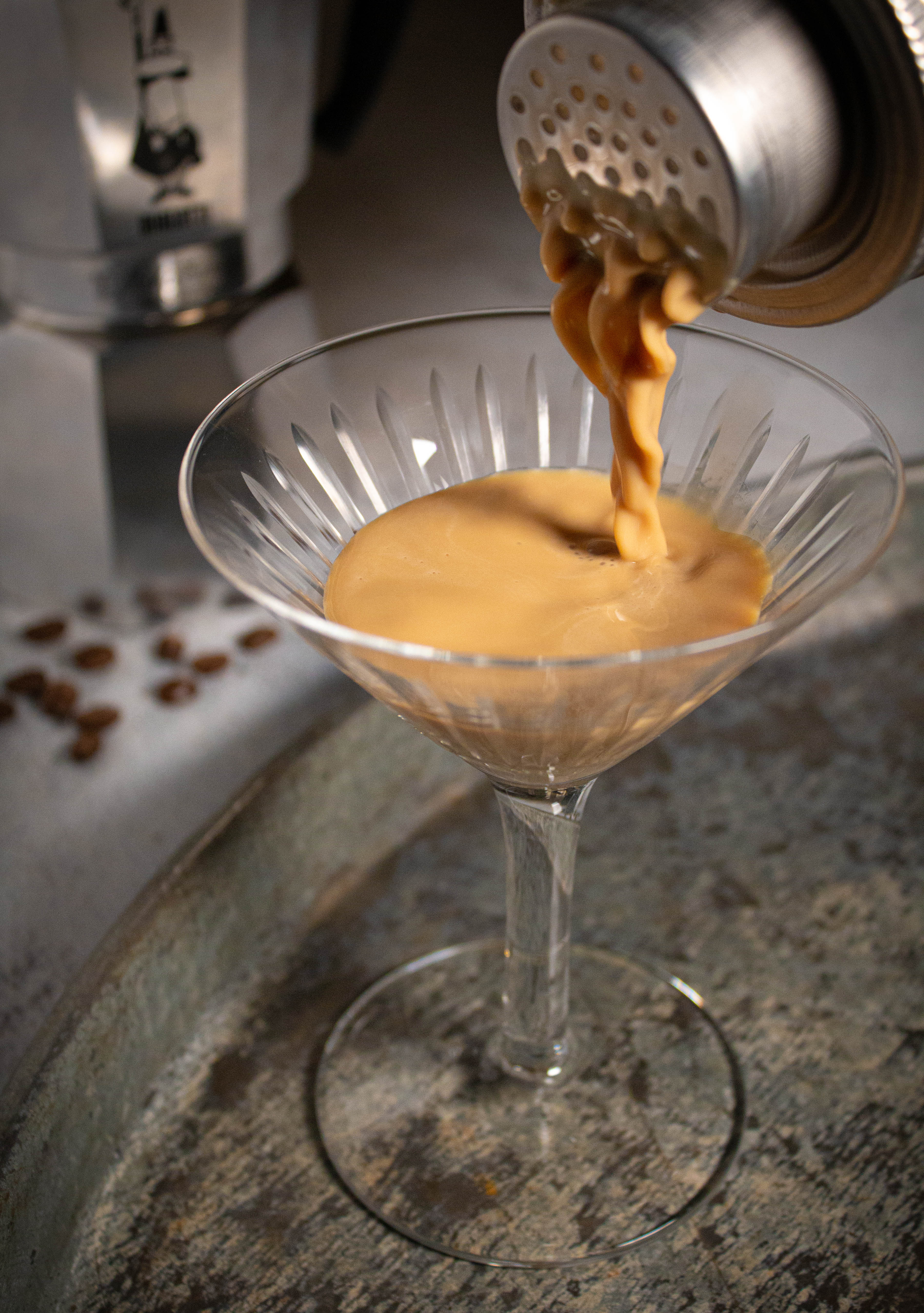 Espresso martini pouring out of brushed stainless steel cocktail shaker into a martini glass with mocha pot and espresso beans in the background.