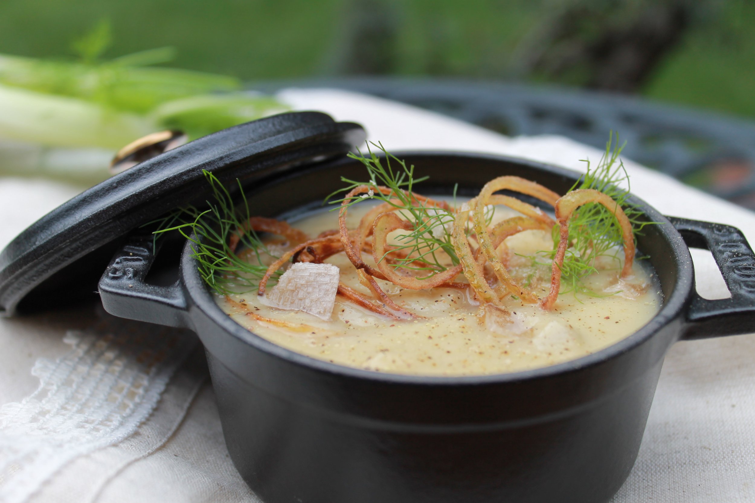 Soup in mini cocotte, garnished with crispy shallots, fennel fronts and flaky sea salt on a beige linen in an outdoor setting.