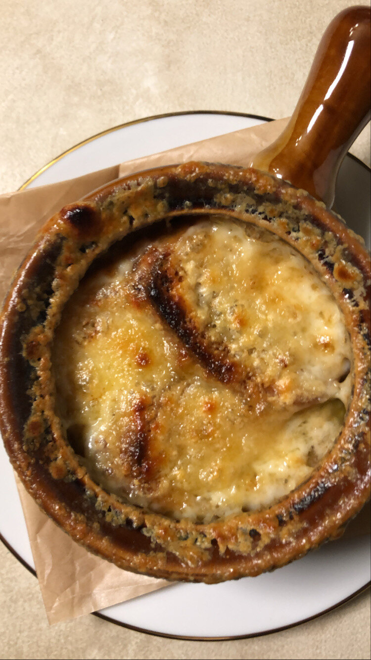 Crock with French onion soup sitting on a parchment square on a gold-rimmed white plate.