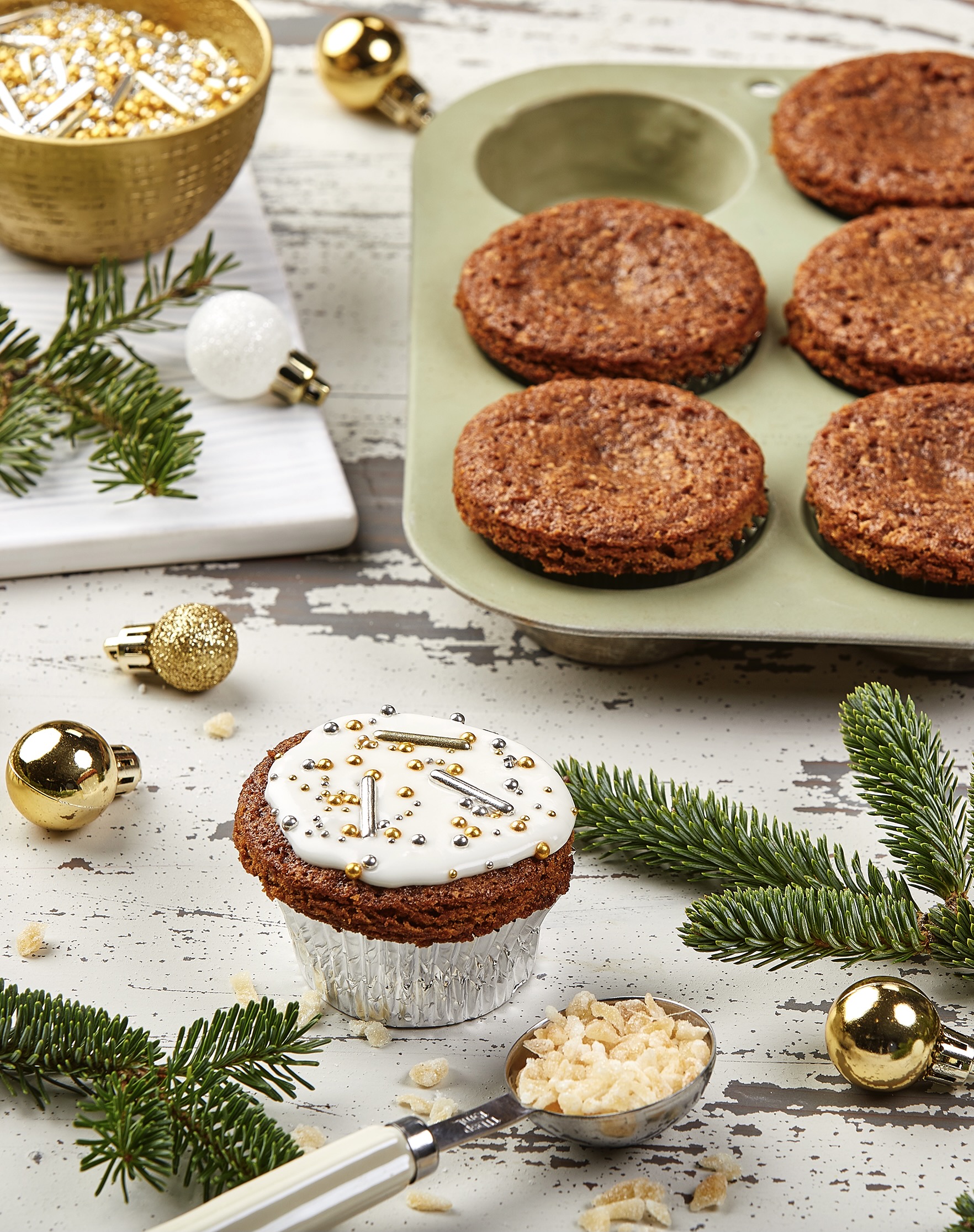 Brown muffin with white glaze and silver and gold sprinkles with evergreen sprigs and mini ornaments in the foreground and background.