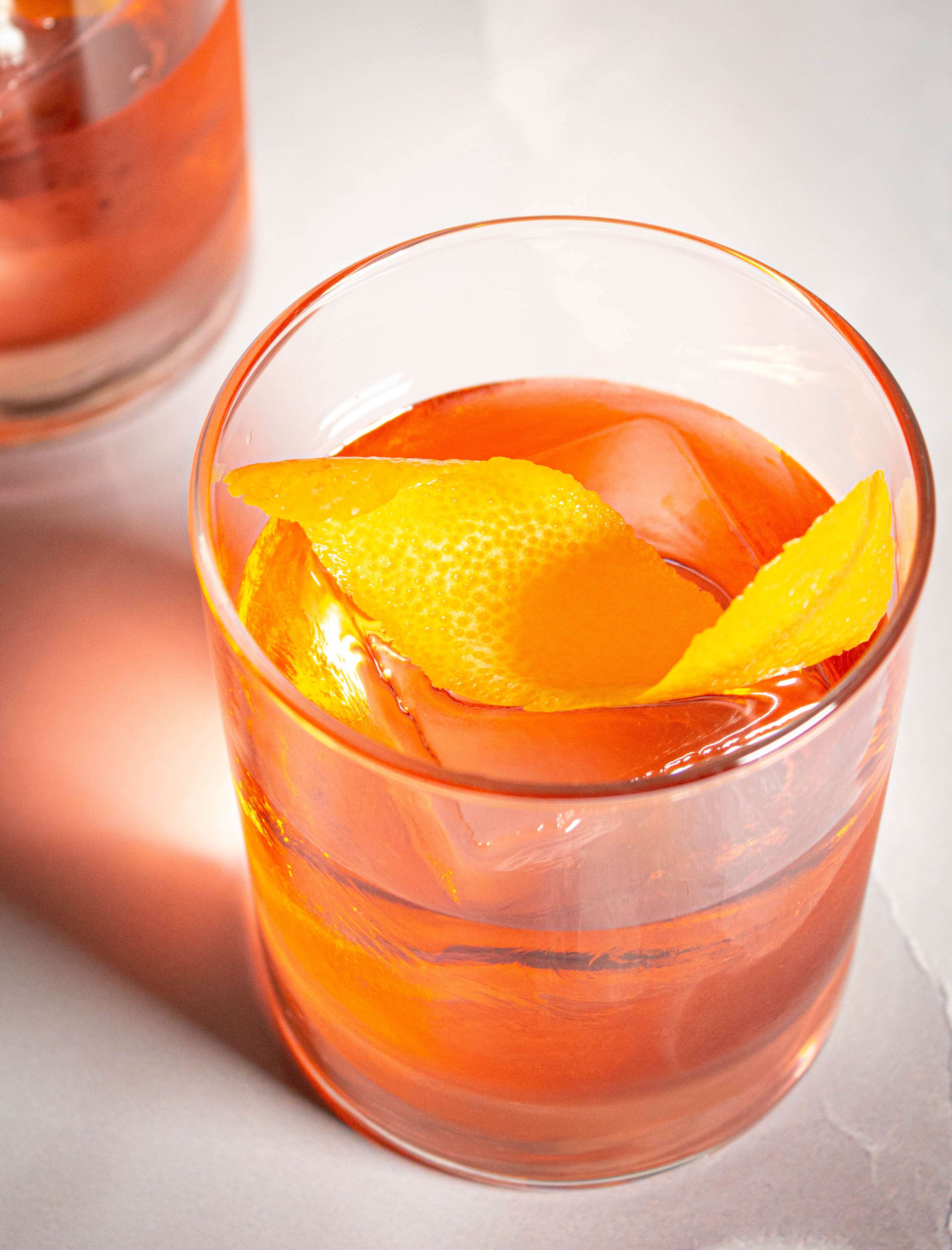 Rocks glass with orange-pink cocktail with a large square cube and orange peel twist.