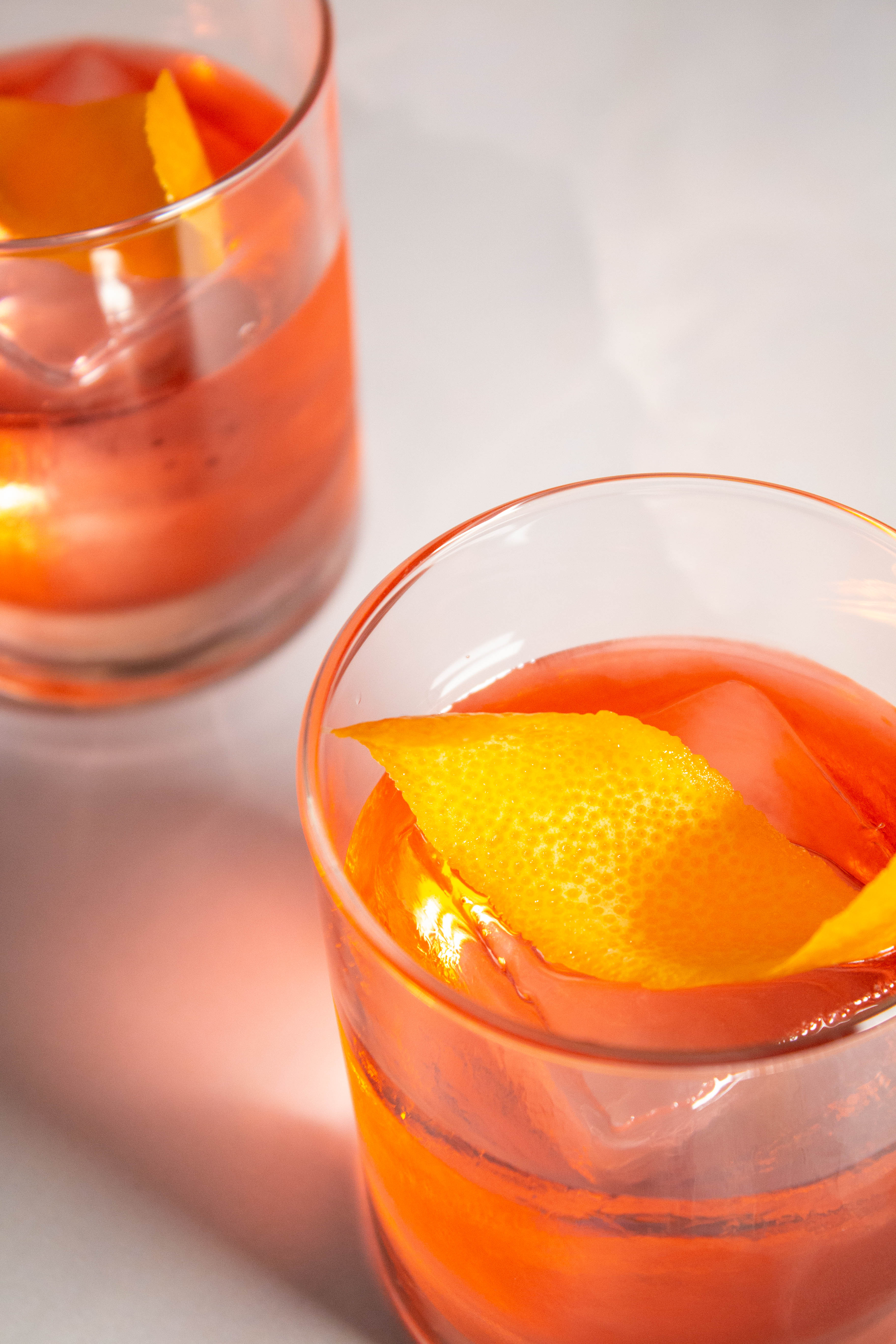 Two rocks glasses with a pinkish-orange cocktail over a large ice cube with an orange peel garnish. 