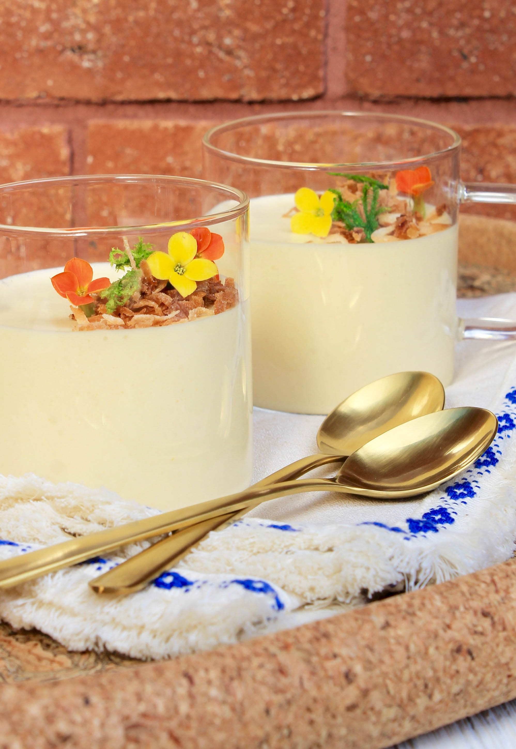 2 mousse cups garnished with toasted coconut and edible flowers on a blue-trimmed napkin with 2 gold spoons and a brick background.