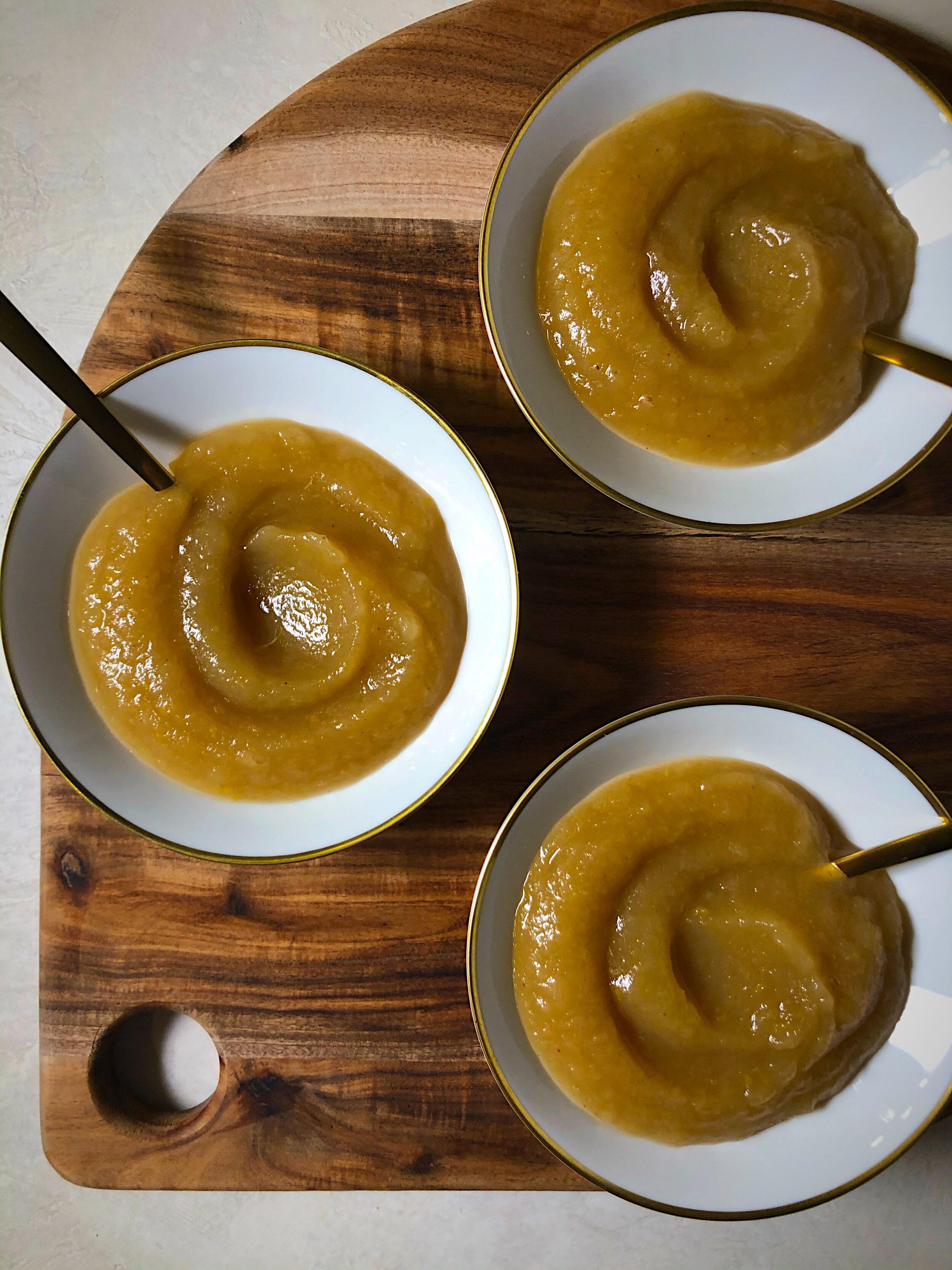 3 bowls with swirls of applesauce on a rounded wooden board.