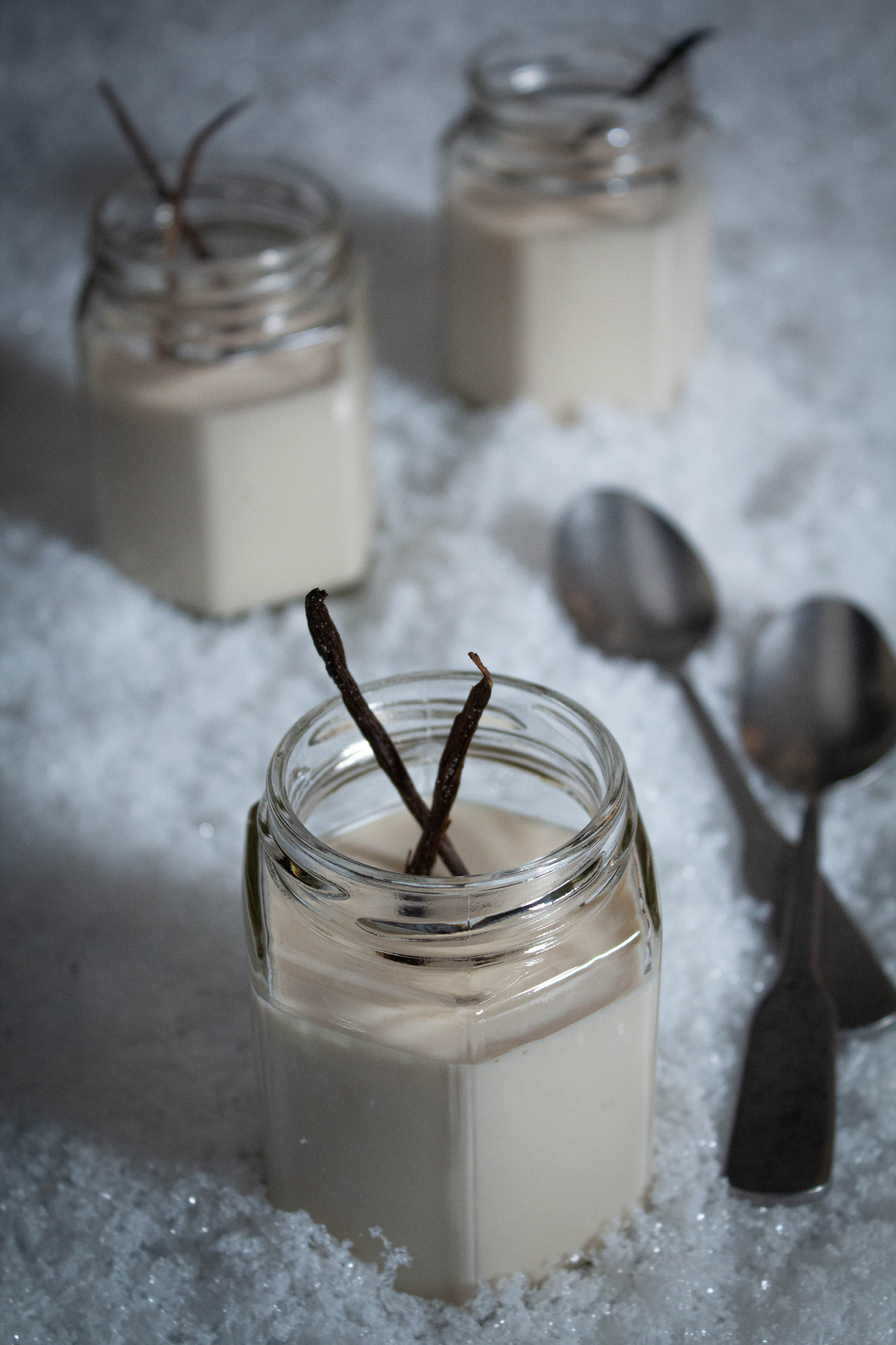 Small glass jars filled with canna cotta garnished with candied vanilla with spoons on a snowy surface.