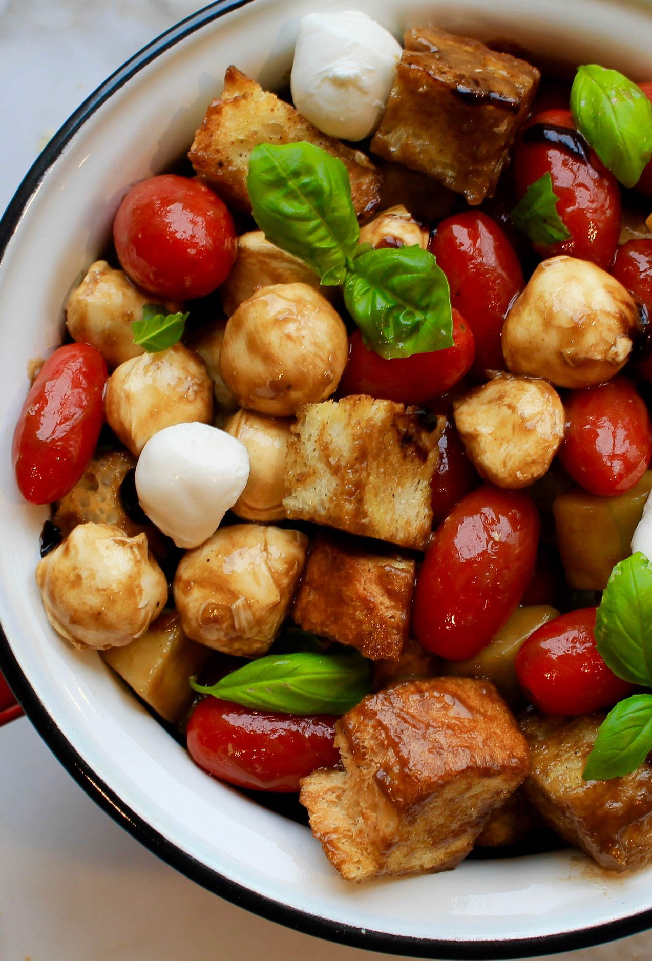 White enameled serving bowl filled with toasted bread, mozzarella balls, cucumber and basil in a balsamic vinaigrette. 