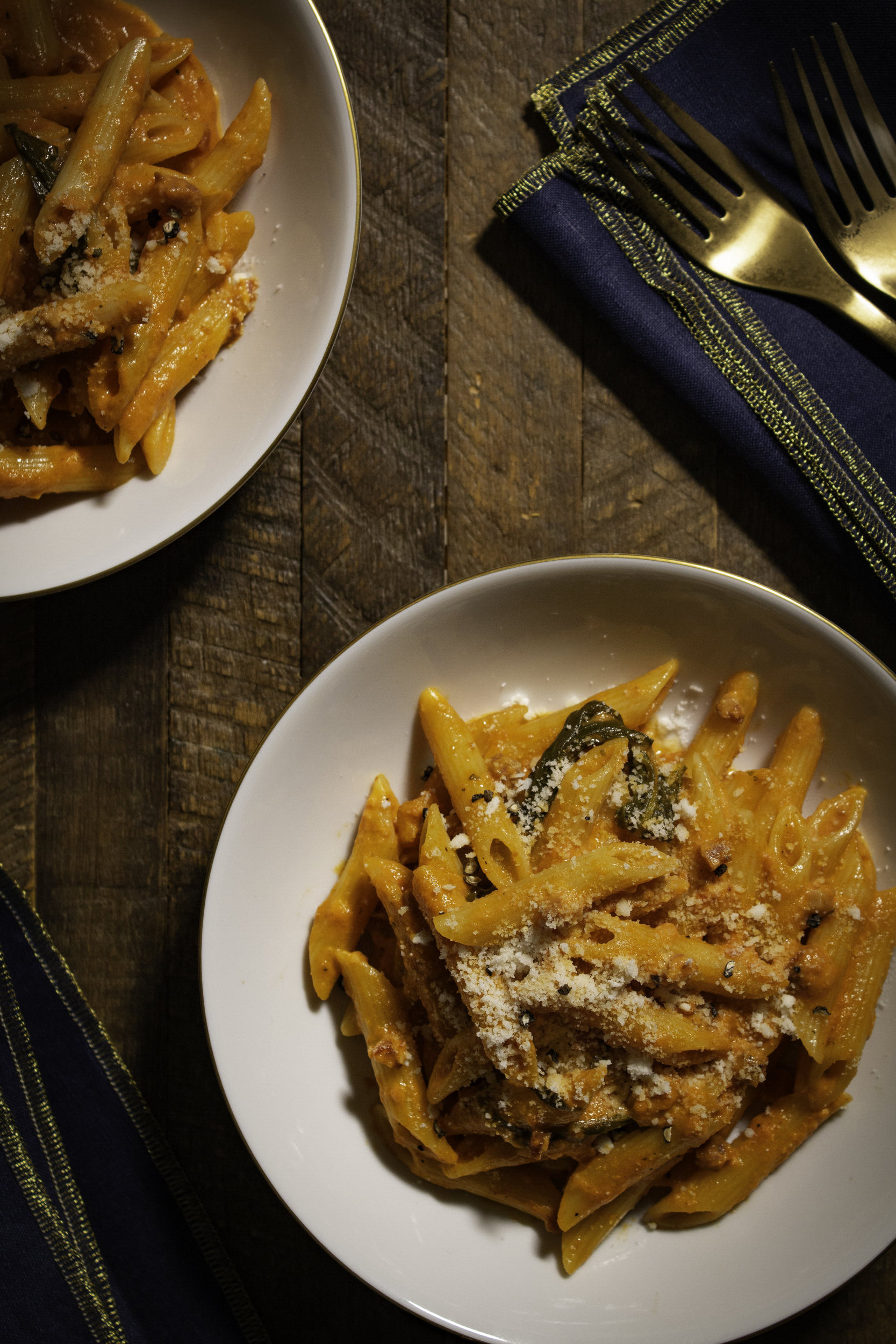 2 plates of penne alla vodka on a dark wooden surface with blue napkins and matte gold forks.