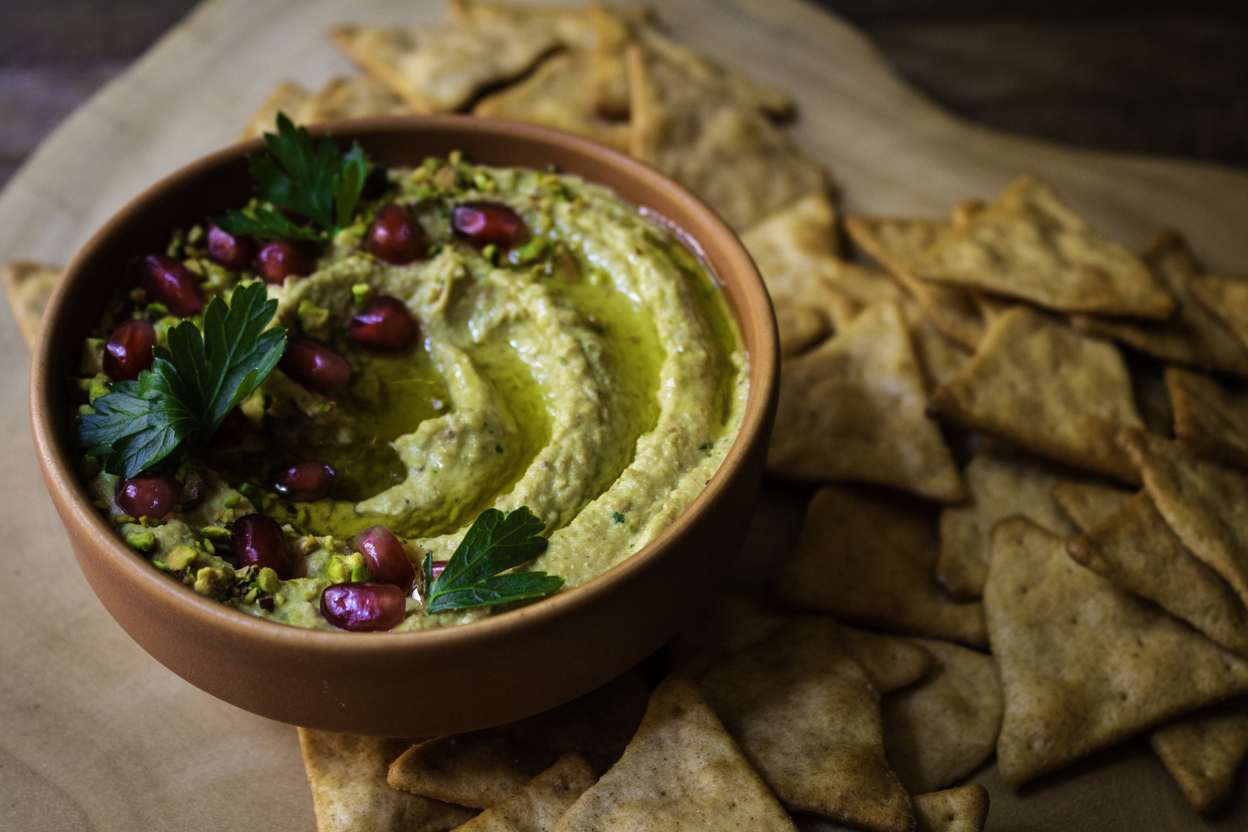Terra cotta-colored bowl with swirls of hummus with parsley, pistachio and pomegranate seed garnish with chips on the side.