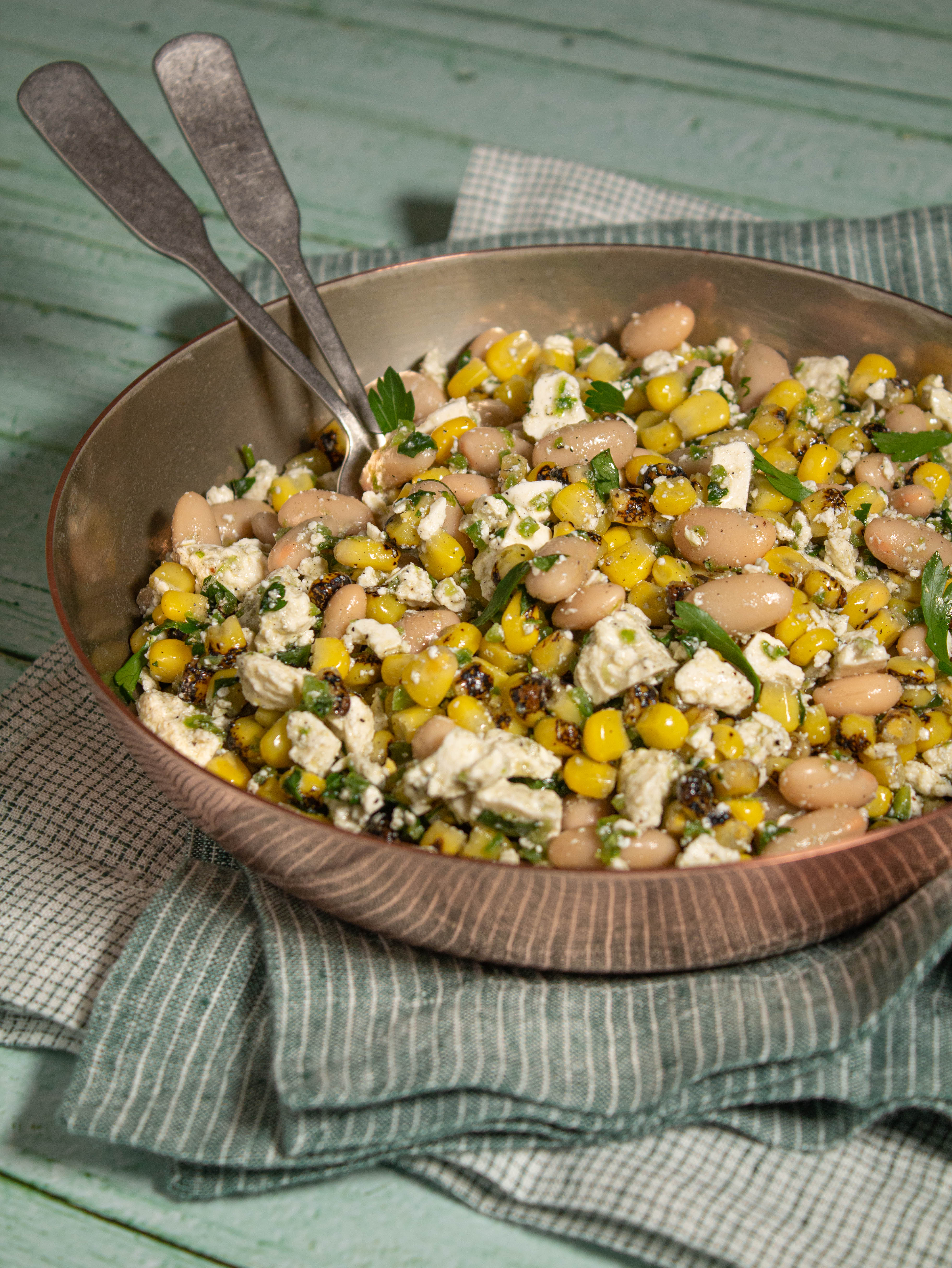Copper pot sitting on green napkins filled with roasted corn, beans, crumbled cheese and cilantro.