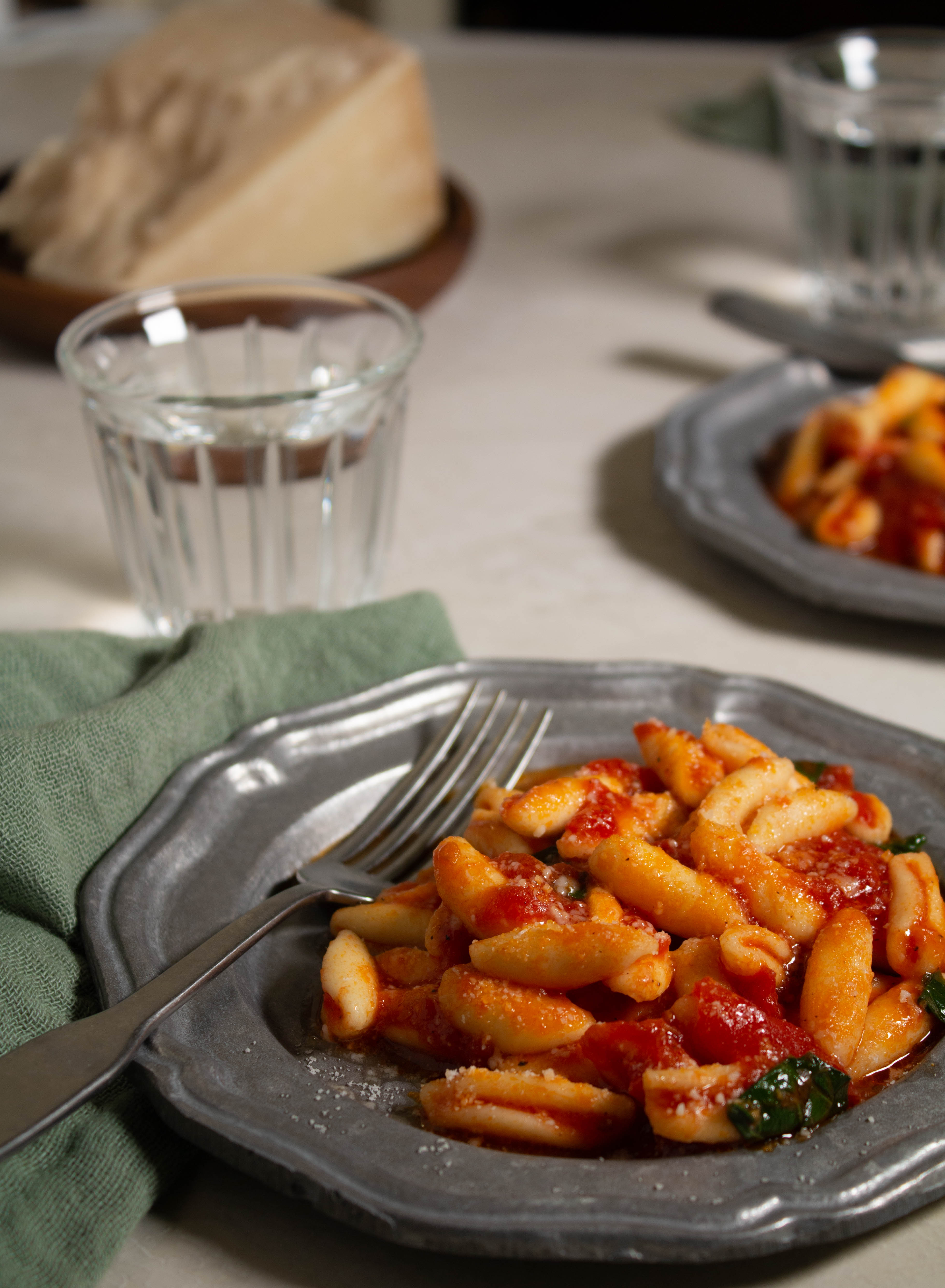 2 pewter plates with cavatelli in tomato sauce with a fork, napkin, water and block of cheese in the background.