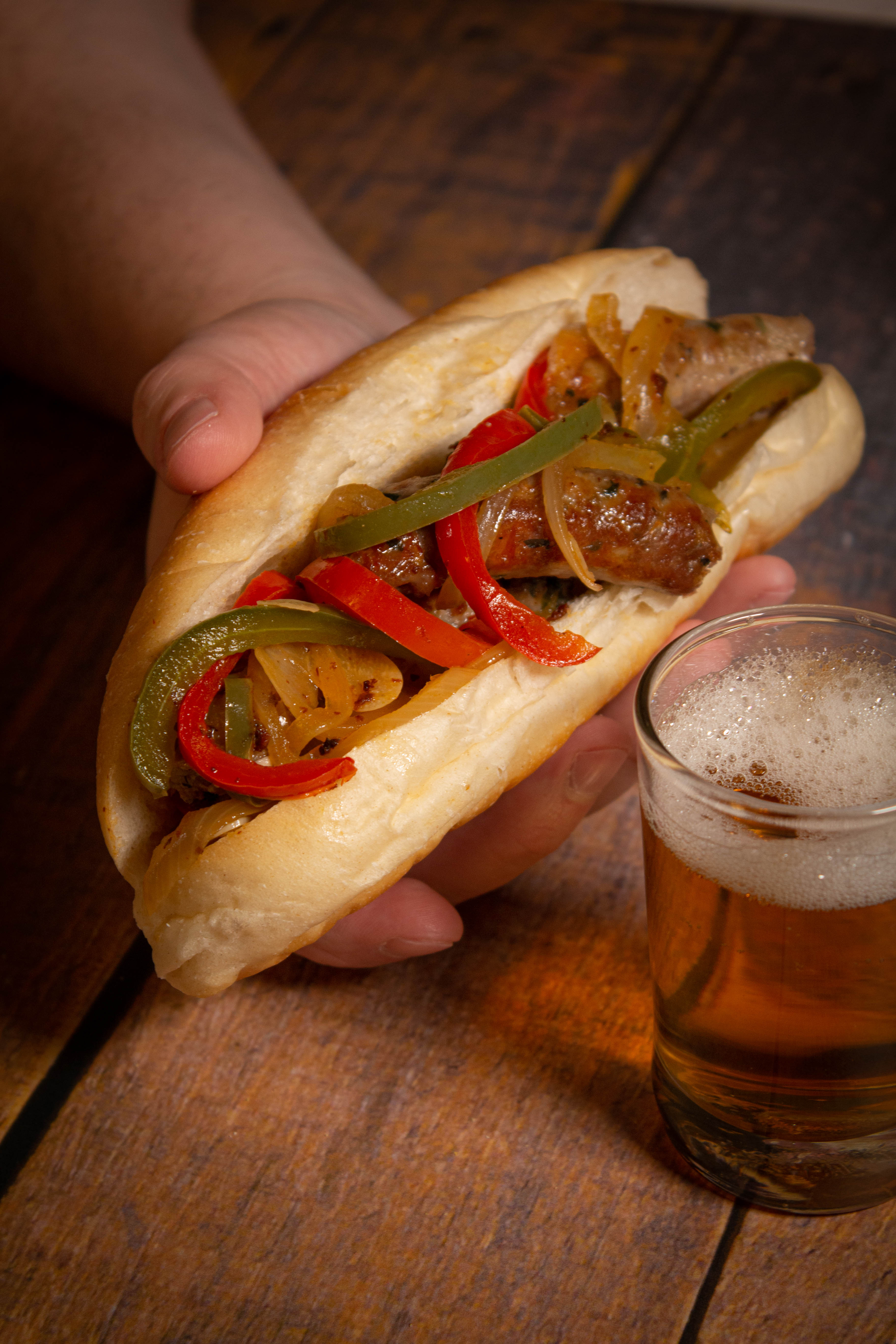 Hand holding a bun filled with sausage and peppers over a wooden surface with a glass of beer in the foreground.