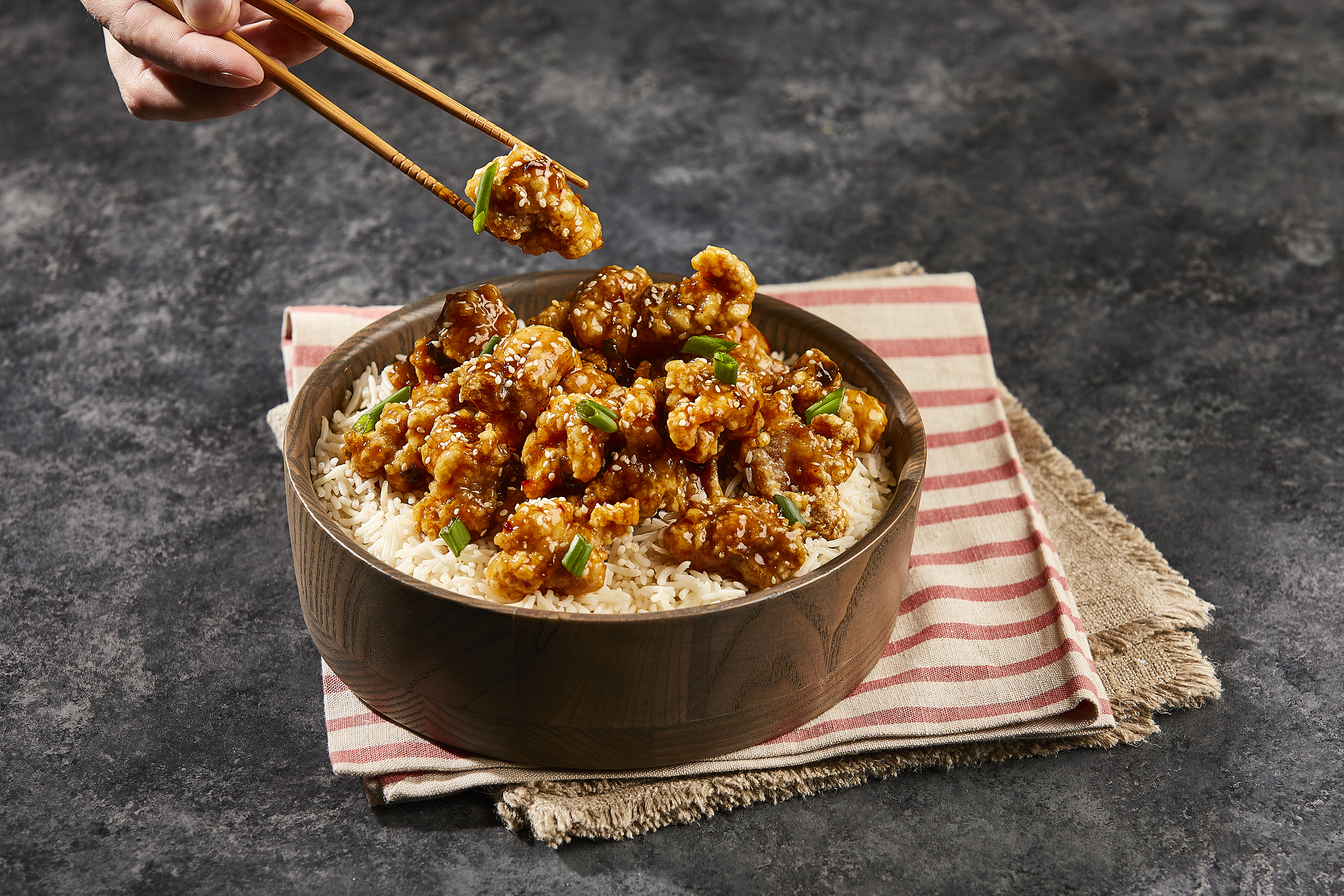 Sesame chicken in a bowl over white rice with a pair of chopsticks lifting one piece out.