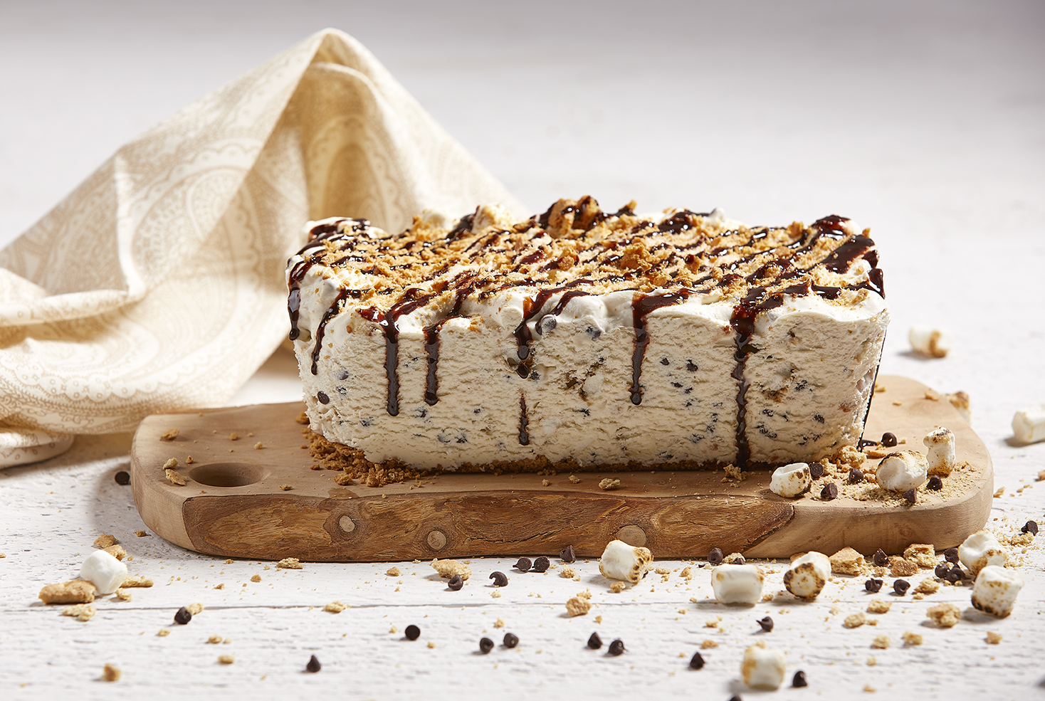 Loaf cake-sized semifreddo topped with toasted marshmallows, graham crumbs and marshmallow sauce on an olive wood board.