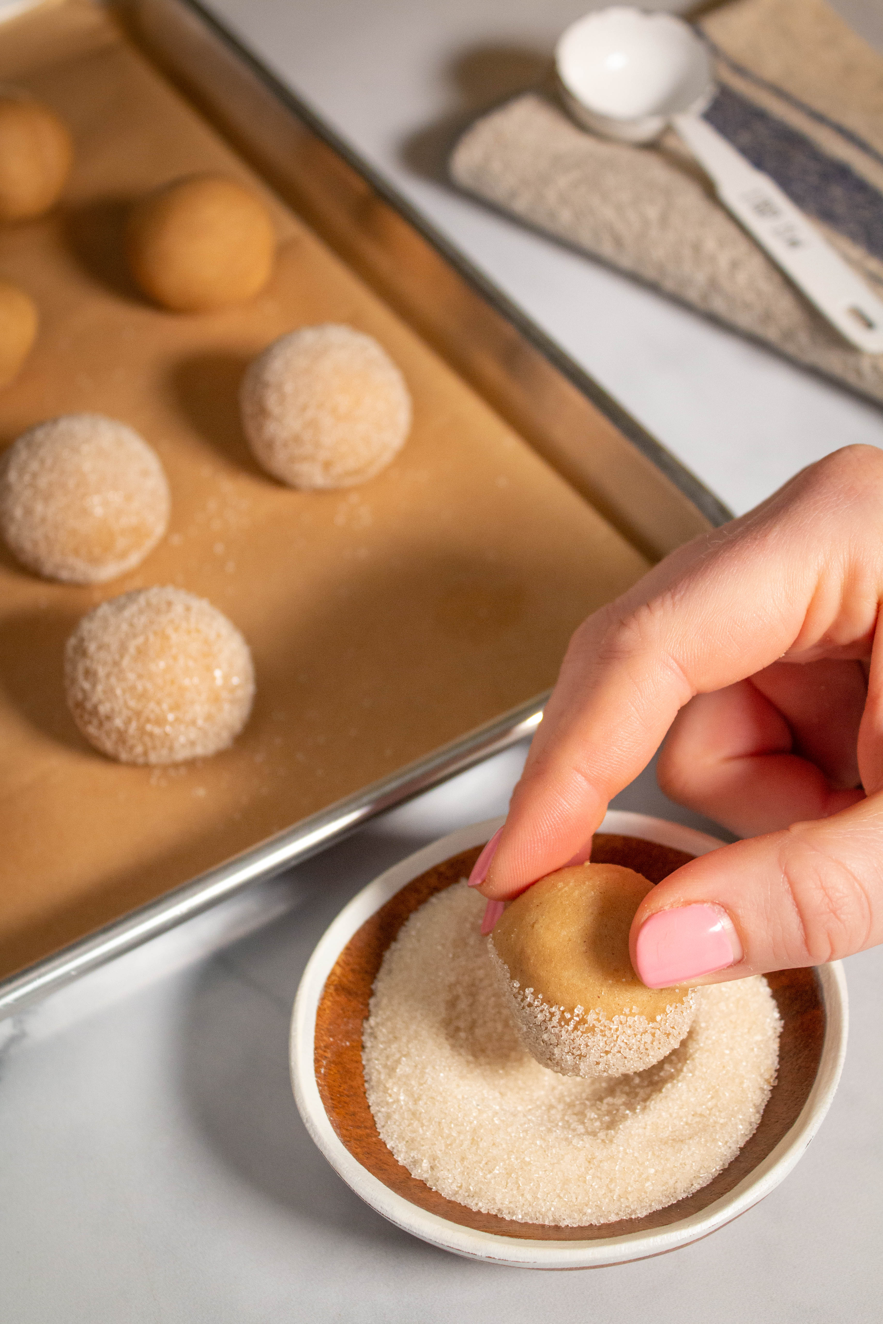 Hand dipping dough ball into sanding sugar with cookie sheet in background.