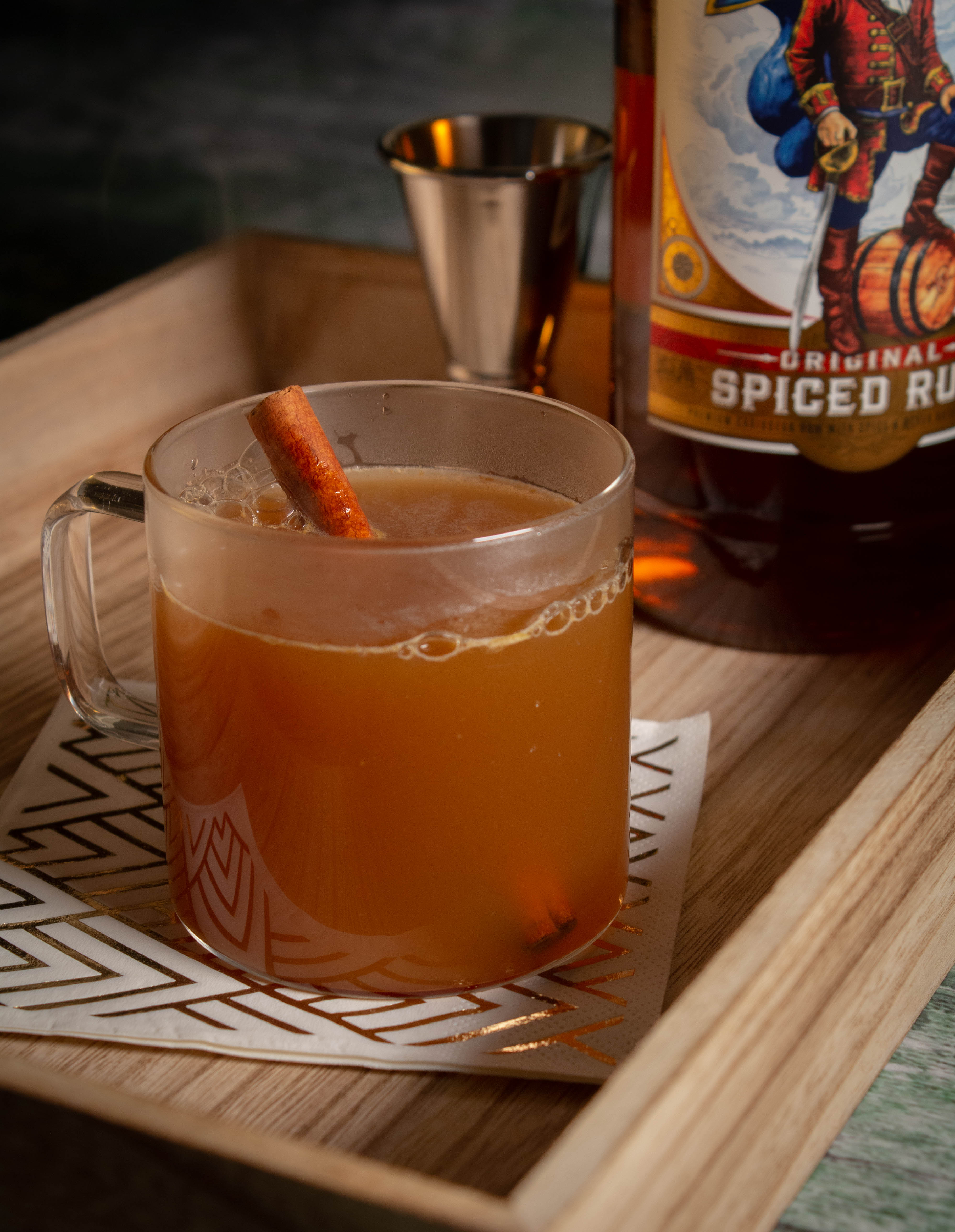 Glass with warm spiced cider and a cinnamon stick with a bottle of spiced rum and a jigger in the background.