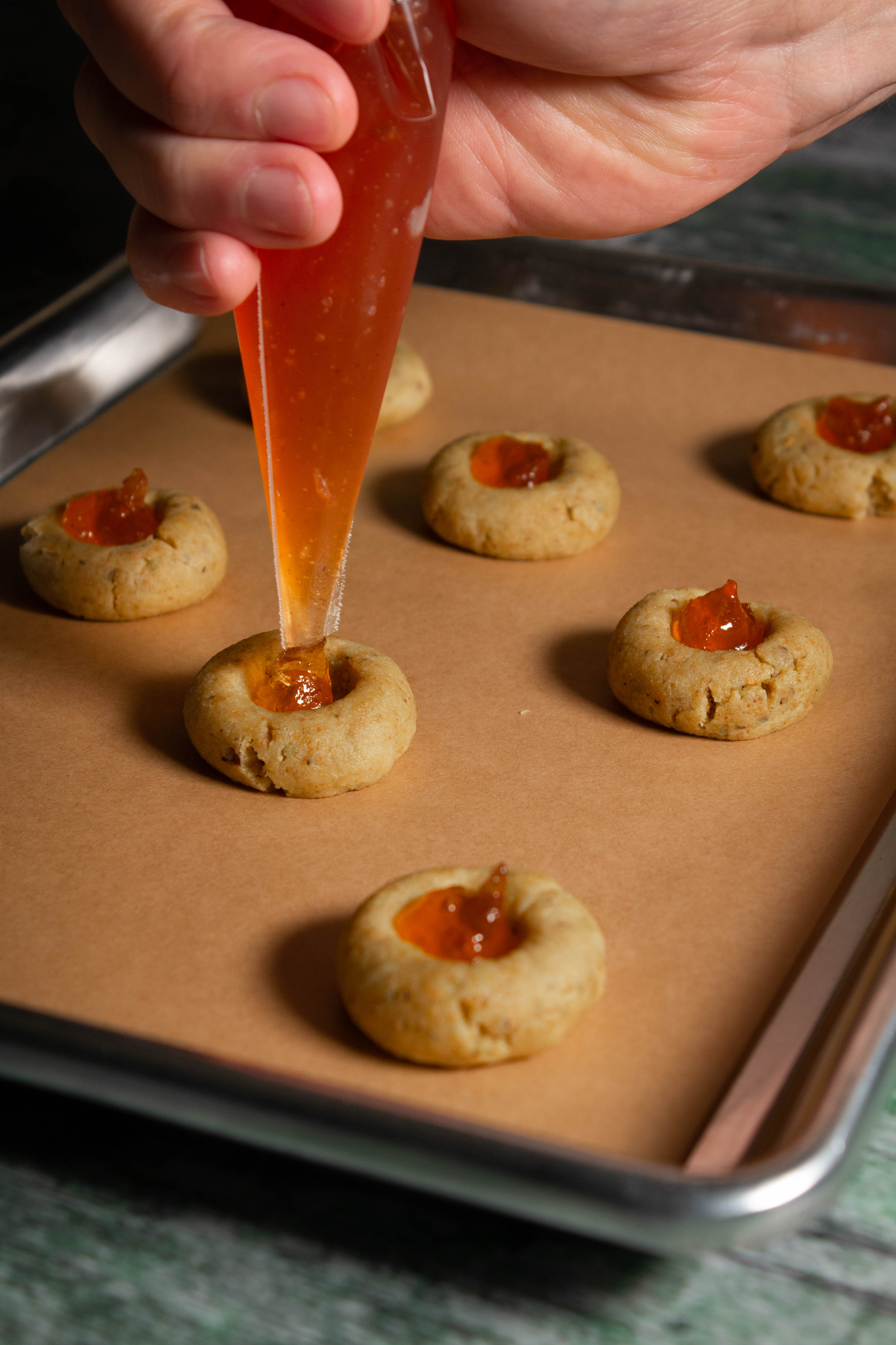 Hand holding a piping bag with apple jam being squeezed onto a cookie on a cookie sheet with others in the background.