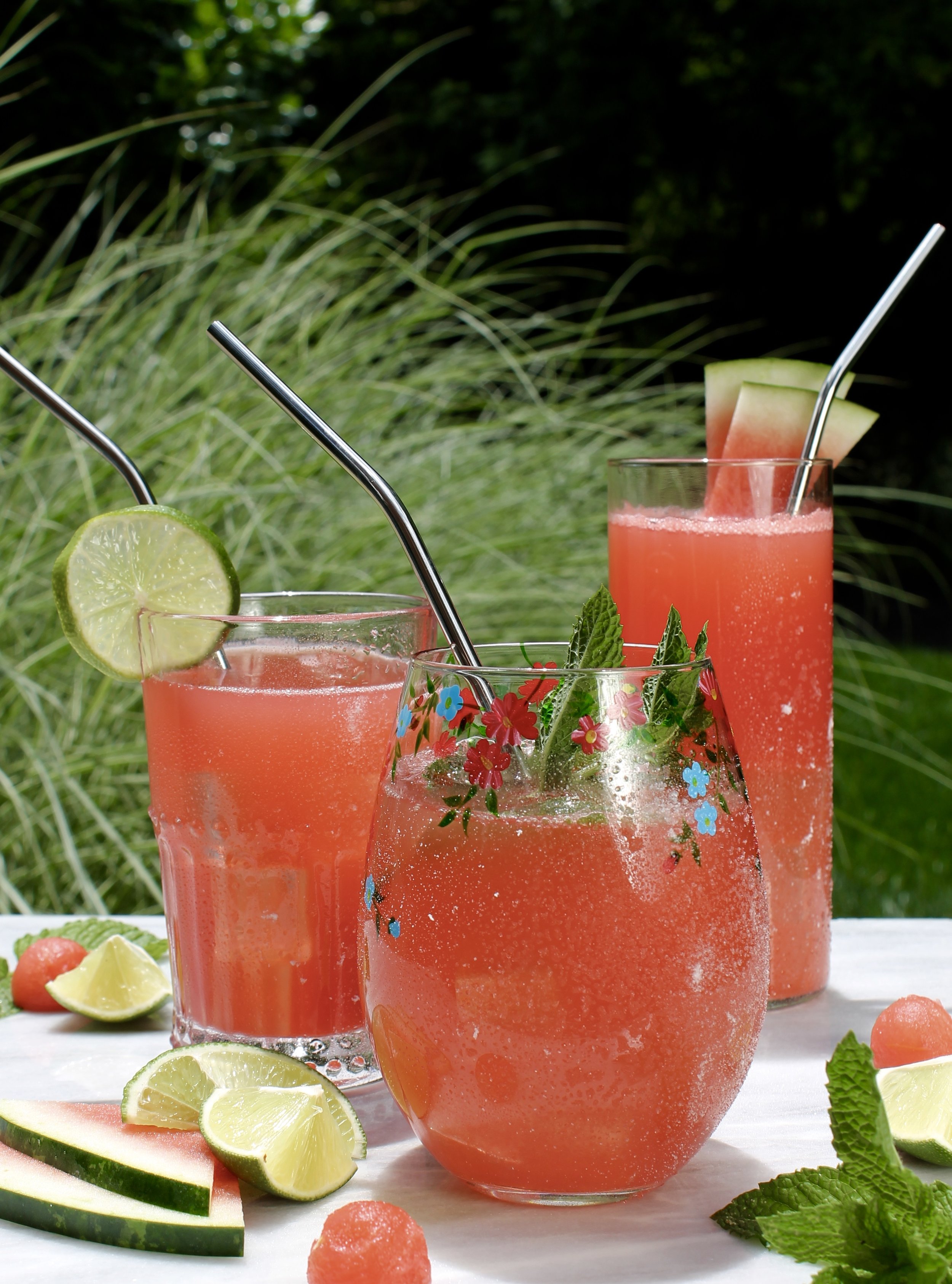 3 glasses filled with a reddish colored cocktail, garnished with mint, lime and watermelon in an outdoor setting.
