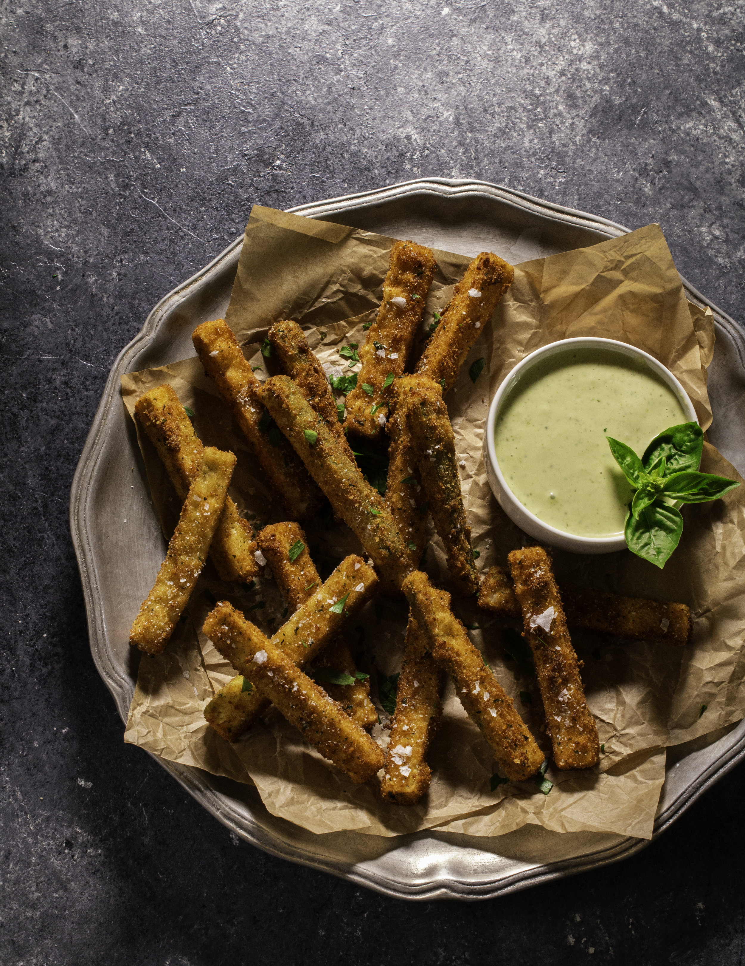 Pewter platter lined with parchment with zucchini fries and a bowl of lemon basil aioli.