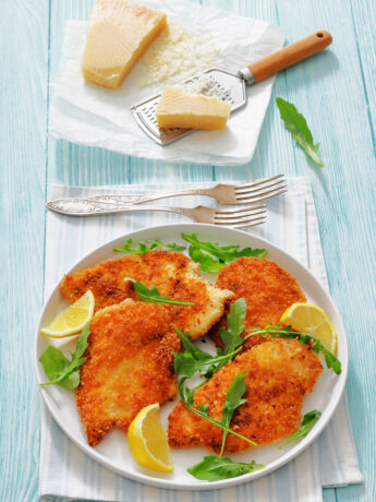 crispy chicken cutlets on a plate with fresh rocket leaves and lemon wedges on a turquoise wooden background with parmesan, grater, forks, vertical view, not AI generated